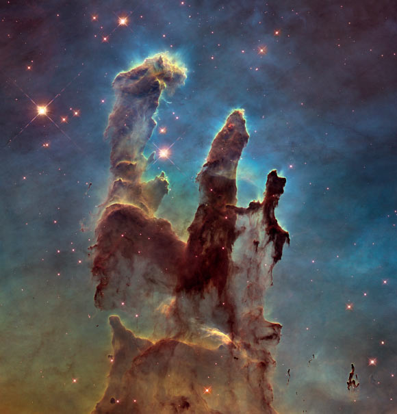 This image shows the Eagle Nebula’s Pillars of Creation as seen in visible light, capturing the multi-colored glow of gas clouds, wispy tendrils of dark cosmic dust, and the rust-colored elephants’ trunks of the nebula’s famous pillars. The dust and gas in the pillars is seared by the intense radiation from young stars and eroded by strong winds from massive nearby stars. Image credit: NASA / ESA / Hubble / Hubble Heritage Team.