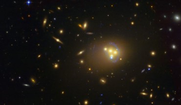 This image shows Abell 3827, a massive galaxy cluster approximately 1.3 billion light years away. The blue structures surrounding the central galaxies are gravitationally lensed views of a much more distant galaxy behind the cluster. Observations of the central four merging galaxies have provided hints that the dark matter around one of the galaxies is not moving with the galaxy itself, possibly implying dark matter-dark matter interactions of an unknown nature are occurring. Image credit: ESO.