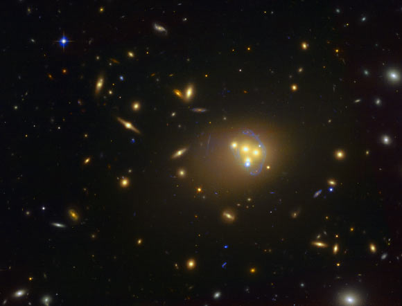 This image shows Abell 3827, a massive galaxy cluster approximately 1.3 billion light years away. The blue structures surrounding the central galaxies are gravitationally lensed views of a much more distant galaxy behind the cluster. Observations of the central four merging galaxies have provided hints that the dark matter around one of the galaxies is not moving with the galaxy itself, possibly implying dark matter-dark matter interactions of an unknown nature are occurring. Image credit: ESO.