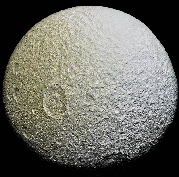This mosaic of Tethys is an orthographic projection constructed from 52 images obtained on April 11, 2015 with Cassini’s narrow-angle camera. The images were obtained at a distance of approximately 33,000 miles (53,000 km) from the moon. The color of the surface can be seen to change across the disk, from yellowish hues to nearly white. These broad color changes are affected by a number of external processes. First, Saturn’s diffuse E-ring preferentially bombards Tethys’ leading hemisphere, toward the right side in this image, with ice bright ice grains. At the same time, charged particles from Saturn’s radiation belt bombard the surface on the trailing side, causing color changes due to chemical alteration of the materials there. The albedo drops by 10 to 15% from the moon’s leading side to the trailing side. Similar global color patterns exist on other Saturnian moons. On a much smaller scale, enigmatic, arc-shaped reddish streaks are faintly visible across the heavily-cratered surface, particularly in the upper right quarter of the image. The origin of this localized color contrast is not yet understood. Image credit: NASA / JPL-Caltech / Space Science Institute.