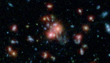 This image, using data from Spitzer and the Hubble Space Telescope, shows the galaxy cluster SpARCS1049+56. At the middle of the picture is the largest, central member of the family of galaxies (upper right red dot of central pair). Unlike other central galaxies in clusters, this one is bursting with the birth of new stars. Image credit: NASA / STScI / ESA / JPL-Caltech / McGill.