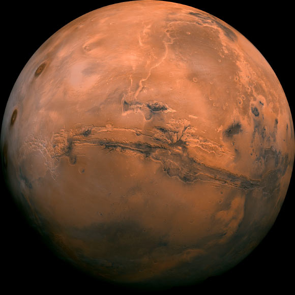 Mosaic of the Valles Marineris hemisphere of Mars projected into point perspective, a view similar to that which one would see from a spacecraft. The distance is 1,550 miles (2,500 km) from the surface of the planet. The mosaic is composed of 102 Viking Orbiter images of Mars. The center of the scene shows the entire Valles Marineris canyon system, over 1,240 miles (2,000 km) long and up to 5 miles (8 km) deep, extending form Noctis Labyrinthus, the arcuate system of graben to the west, to the chaotic terrain to the east. Many huge ancient river channels begin from the chaotic terrain from north-central canyons and run north. The three Tharsis volcanoes (dark red spots), each about 15.5 miles (25 km) high, are visible to the west. South of Valles Marineris is very ancient terrain covered by many impact craters. Image credit: NASA / JPL-Caltech.