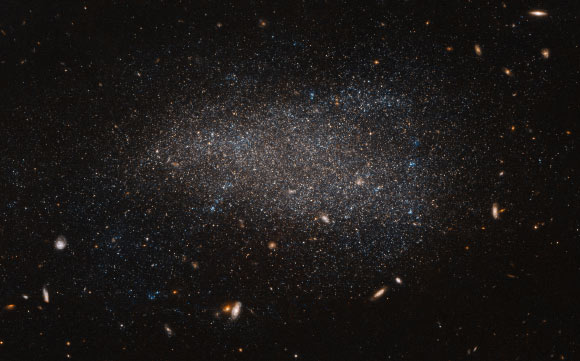 This Hubble image shows the dwarf irregular galaxy NGC 4789A. The image also reveals numerous other galaxies, far more distant, that appear as fuzzy shapes spread across the image. The colors in this image have been deliberately exaggerated to emphasize the mix of blue and red stars. The blue stars are bright, hot and massive stars that have formed relatively recently, whereas the red stars are much older. The presence of both tells us that stars have been forming in this galaxy throughout its history. Image credit: NASA / ESA / Hubble / Judy Schmidt, www.geckzilla.com.