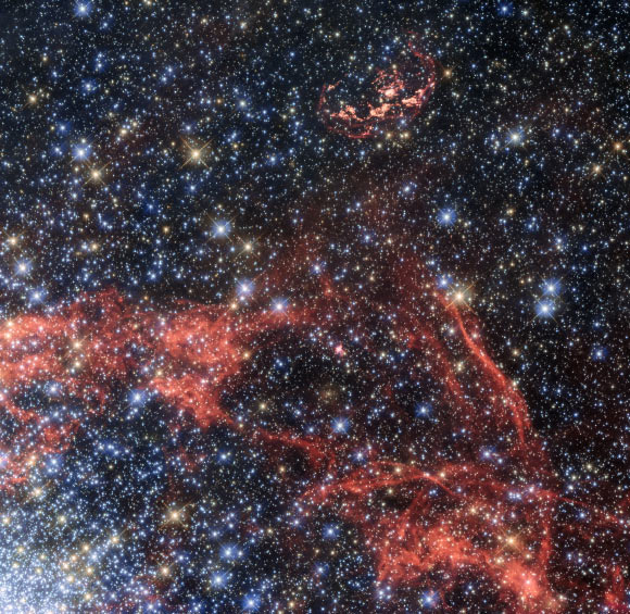 This Hubble image shows the supernova remnant N103B (top of the image). N103B was a Type Ia supernova, located in the Large Magellanic Cloud. Owing to its relative proximity to Earth, astronomers observe the remnant to search for a potential stellar survivor of the explosion. The orange-red filaments visible in the image show the shock fronts of the supernova explosion. These filaments allow astronomers to calculate the original centre of the explosion. The filaments also show that the explosion is no longer expanding as a sphere, but is elliptical in shape. Astronomers assume that part of material ejected by the explosion hit a denser cloud of interstellar material, which slowed its speed. The shell of expanding material being open to one side supports this idea. The gas in the lower half of the image and the dense concentration of stars in the lower left are the outskirts of the rich star cluster NGC 1850. Image credit: NASA / ESA / Hubble.
