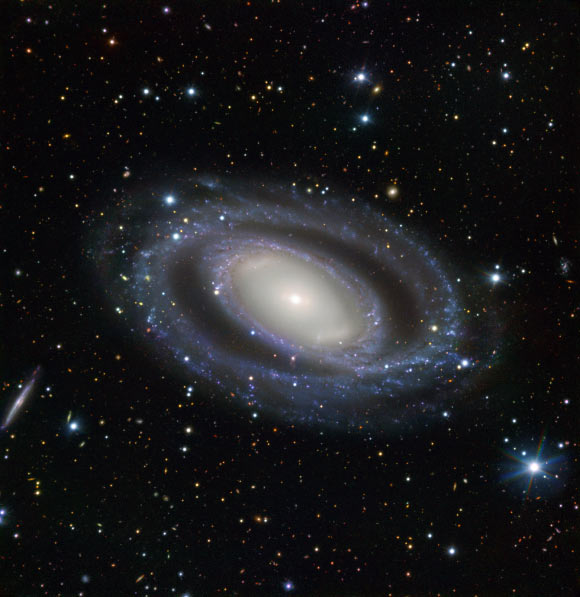 This image shows the barred-ring spiral galaxy NGC 7098 (center). An array of distant galaxies are also visible throughout the frame, the most prominent being the small, edge-on, spiral galaxy visible to the left of NGC 7098, known as ESO 048-G007. Image credit: ESO.