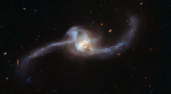 NGC 2623 is in the late stages of the merging process, with the centers of the original galaxy pair now merged into one nucleus, but stretching out from the center are two tidal tails of young stars, a strong indicator that a merger has taken place. During such a collision, the dramatic exchange of mass and gases initiates star formation, seen here in both the tails. The prominent lower tail is richly populated with bright star clusters. These star clusters may have formed as part of a loop of stretched material associated with the northern tail, or they may have formed from debris falling back onto the nucleus. In addition to this active star-forming region, both galactic arms harbor very young stars in the early stages of their evolutionary journey. Image credit: NASA / ESA / Hubble.