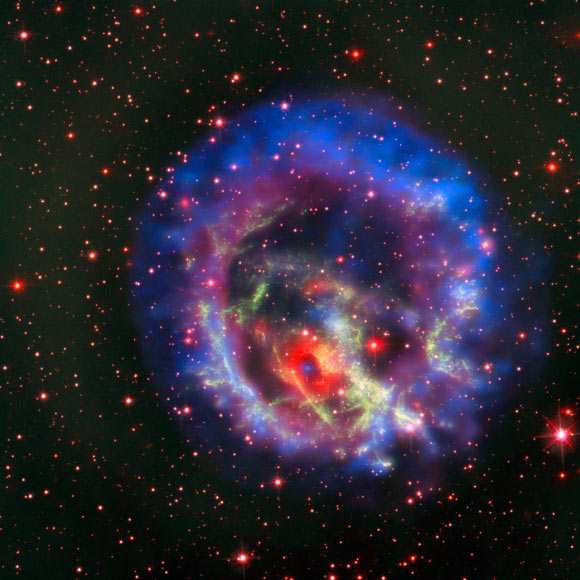 This composite picture created from images from several space- and ground-based telescopes tells the story of the hunt for an elusive missing object hidden amid a complex tangle of gaseous filaments in the Small Magellanic Cloud, a dwarf galaxy approximately 200,000 light-years from us outside the Milky Way Galaxy. The reddish background image comes from the NASA/ESA Hubble Space Telescope and reveals the wisps of gas forming the supernova remnant 1E 0102.2-7219 in green. The red ring with a dark center is from the MUSE instrument on ESO’s Very Large Telescope and the blue and purple images are from NASA’s Chandra X-Ray Observatory. The blue spot at the center of the red ring is an isolated neutron star with a weak magnetic field, the first identified outside the Milky Way. Image credit: ESO / NASA / ESA / Hubble Heritage Team / STScI / AURA / F. Vogt et al.