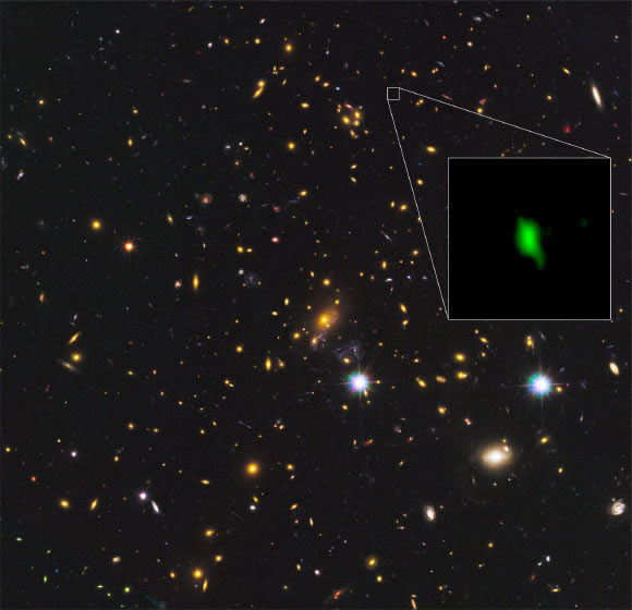 This image shows the galaxy cluster MACS J1149.5+2223 taken with the NASA/ESA Hubble Space Telescope; the inset image is the gravitationally lensed galaxy MACS1149-JD1, seen as it was 13.28 billion years ago and observed with ALMA. Here, the oxygen distribution detected with ALMA is depicted in green. Image credit: ALMA / ESO / NAOJ / NRAO / NASA / ESA / Hubble Team / W. Zheng, JHU / M. Postman, STScI / CLASH Team / Hashimoto et al.