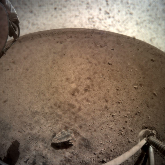 InSight flipped open the lens cover on its Instrument Context Camera (ICC) on November 30, 2018, and captured this view of Mars. Located below the deck of the InSight lander, the ICC has a fisheye view, creating a curved horizon. Some clumps of dust are still visible on the camera’s lens. One of the spacecraft’s footpads can be seen in the lower right corner. The seismometer’s tether box is in the upper left corner. Image credit: NASA / JPL-Caltech.