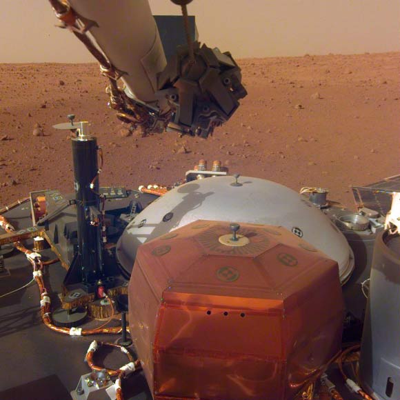 This image from InSight’s robotic-arm mounted Instrument Deployment Camera shows the instruments on the spacecraft’s deck, with the Martian surface of Elysium Planitia in the background. The color-calibrated picture was acquired on December 4, 2018 (Sol 8); in the foreground, a copper-colored hexagonal cover protects the SEIS seismometer; the gray dome behind SEIS is the wind and thermal shield, which will be placed over SEIS; to the left is a black cylindrical instrument, the Heat Flow and Physical Properties Probe (HP3); HP3 will drill up to 16 feet (5 m) below the Martian surface, measuring heat released from the interior of the planet; above the deck is InSight’s robotic arm, with the stowed grapple directly facing the camera; to the right can be seen a small portion of one of the two solar panels that help power InSight and part of the UHF communication antenna. Image credit: NASA / JPL-Caltech.