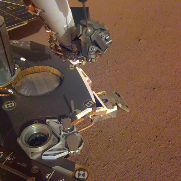 A partial view of InSight’s deck, where it stands on the Martian plains Elysium Planitia; the color-calibrated image was received on December 4, 2018 (Sol 8); InSight’s robotic arm with its stowed grapple can be seen above the deck, and jutting out from the front of the deck is one of the boxy attitude control system thrusters that helped control the spacecraft’s landing; the circular silver inset of the propellant tank can also be seen in the middle of the image, as well as one of the connections for the aeroshell and parachute, which looks like a cupholder in the foreground; next to the propellant tank is the UHF antenna, which helps the lander communicate with Earth; in the background, part of one of InSight’s solar panels is visible. Image credit: NASA / JPL-Caltech.