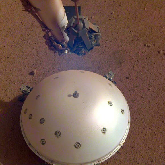 This image, taken March 19, 2019 by a camera on NASA’s InSight lander, shows its Wind and Thermal Shield, which covers its seismometer, the SEIS, and the Martian surface in the background. Image credit: NASA / JPL-Caltech.
