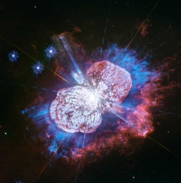 Astronomers have monitored the Eta Carinae binary system for more than two decades. It has been prone to violent outbursts, including an episode in the 1840s during which ejected material formed the bipolar bubbles seen here. Now, using Hubble’s Wide Field Camera 3 (WFC3) instrument to probe the nebula in ultraviolet light, they have uncovered the glow of magnesium embedded in warm gas (shown in blue) in places they had not seen it before. The luminous magnesium resides in the space between the dusty bipolar bubbles and the outer shock-heated nitrogen-rich filaments (shown in red). The streaks visible in the blue region outside the lower-left lobe are a striking feature of the image. These streaks are created when the star’s light rays poke through the dust clumps scattered along the bubble’s surface. Wherever the ultraviolet light strikes the dense dust, it leaves a long, thin shadow that extends beyond the lobe into the surrounding gas. Image credit: NASA / ESA / Hubble / N. Smith, University of Arizona / J. Morse, BoldlyGo Institute.