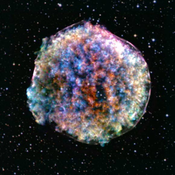 This image of Tycho’s SNR from NASA’s Chandra X-ray Observatory reveals an intriguing pattern of bright clumps and fainter holes. To emphasize these features, two narrow ranges of X-ray energies were selected to isolate material (silicon, colored red) moving away from Earth, and moving towards us (also silicon, colored blue). The other colors in the image (yellow, green, blue-green, orange and purple) show a broad range of different energies and elements, and a mixture of directions of motion. Chandra’s X-ray data are shown alongside an optical image of the stars in the same field of view from the Digitized Sky Survey. Image credit: NASA / CXC / RIKEN / NASA’s Goddard Space Flight Center / T. Sato et al / DSS.