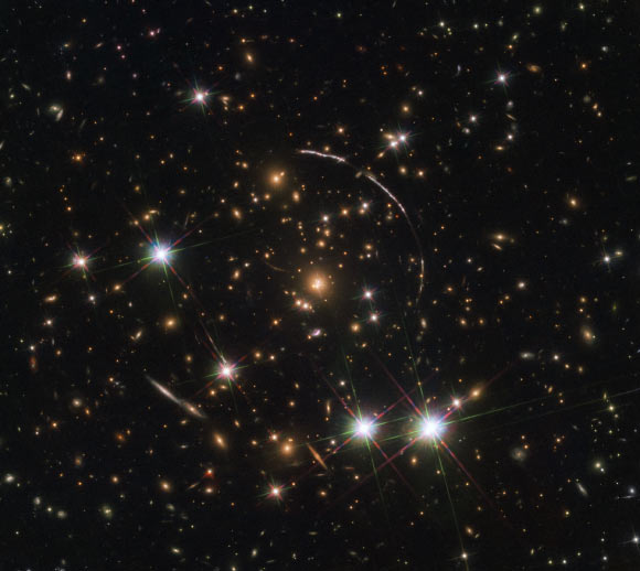 This Hubble image shows a massive galaxy cluster, about 4.6 billion light years away. Along its borders 12 images within the four arcs are visible; these are copies of the same galaxy called PSZ1 G311.65-18.48, which is almost 11 billion light-years away. Three of these arcs are visible in the top right of the image, while one counterarc is visible in the lower left -- partially obscured by a bright foreground star within our Milky Way Galaxy. Image credit: NASA / ESA / Hubble / Rivera-Thorsen et al.