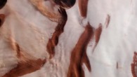 This HRSC image shows plumes of dust clouds over the ice cap at Mars’ north pole. The landscape here is a rippled mix of color. Dark red and ochre-hued troughs appear to cut through the icy white of the polar cap; these form part of a wider system of depressions that spiral outwards from the very center of the pole. Visible to the left of the frame are a few extended streams of clouds, aligned perpendicularly to a couple of the troughs. These are thought to be caused by small local storms that kick up dust into the Martian atmosphere, eroding scarps and slopes as they do so and slowly changing the appearance of the troughs over time. Image credit: ESA / DLR / FU Berlin / CC BY-SA 3.0 IGO.