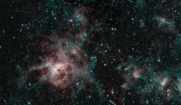 This image of the Tarantula Nebula combines data from multiple Spitzer observations, most recently in February and September 2019. The image shows the nebula in two wavelengths of infrared light, each represented by a different color. The red color at the heart of the nebula shows the presence of particularly hot gas emitting infrared light at a wavelength of 4.5 micrometers. The blue regions are dust composed of molecules called polycyclic aromatic hydrocarbons. Regions emitting both wavelengths appear white. Image credit: NASA / JPL-Caltech.