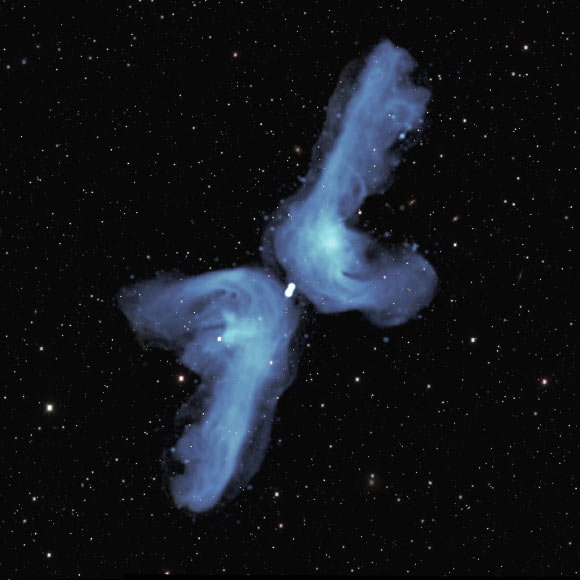 PKS 2014-55 is classified as ‘X-shaped’ because of its appearance in previous relatively blurry images. The detail provided in this radio image obtained with the MeerKAT telescope indicates that its shape is best described as a double boomerang. Two powerful jets of radio waves, indicated in blue color, each extend 2.5 million light-years into space. Eventually, they are ‘turned back’ by the pressure of tenuous intergalactic gas. As they flow back towards the central galaxy, they are deflected by its relatively high gas pressure into the shorter, horizontal, arms of the boomerang. The background image shows visible light from myriad galaxies in the distant Universe. Image credit: NRAO / AUI / NSF / SARAO / DES.