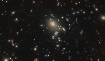 This Hubble image shows a massive galaxy cluster (center); along its borders reddish dots are visible; these are copies of the same galaxy called PLCK G045.1+61.1, which is 12 billion light-years away. Image credit: NASA / ESA / Hubble / B. Frye.