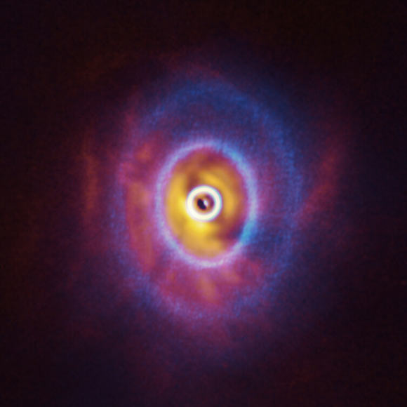 This composite image shows both ALMA and SPHERE/VLT observations of the disk around GW Orionis. The ALMA image (blue) shows the disk’s ringed structure, with the innermost ring (part of which is visible as an oblong dot at the very center of the image) separated from the rest of the disk. The SPHERE observations (orange-red) allowed the astronomers to see for the first time the shadow of this innermost ring on the rest of the disk, which made it possible for them to reconstruct its warped shape. Image credit: ALMA / ESO / NAOJ / NRAO / University of Exeter / Kraus et al.