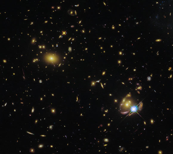 This Hubble image shows a massive galaxy cluster (bottom left corner) and three images of the gravitationally lensed galaxy SGAS 0033+02. The color image was made from separate exposures taken in the visible and infrared regions of the spectrum with Hubble’s Wide Field Camera 3 (WFC3). Five filters were used to sample various wavelengths. The color results from assigning different hues to each monochromatic image associated with an individual filter. Image credit: NASA / ESA / Hubble / E. Wuyts.