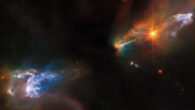 Two wispy, gaseous clouds occupy the corners of this image, HH 1 in the upper right, and HH 2 in the lower left. Both are light blue and surrounded by dimmer multi-colored clouds, while the background is dark black due to dense gas. A very bright orange star lies just to the lower left of HH 1, and beyond that star is a narrow jet, emerging from the dark center of the field. Image credit: NASA / ESA / Hubble / B. Reipurth / B. Nisini.