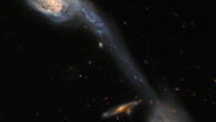 This image shows two member galaxies, PGC 36733 (left) and PGC 36723 (right), in the Wild’s Triplet. The galaxies are a mix of pale blue and yellow in color, crossed by strands of dark red dust. A long, faint streak of pale blue joins them, extending from an arm of one galaxy and crossing the field diagonally. A small spiral galaxy, orange in color, is visible edge-on, left of PGC 36723. Image credit: NASA / ESA / Hubble / Dark Energy Survey / DOE / FNAL / DECam / CTIO / NOIRLab / NSF / AURA / J. Dalcanton.