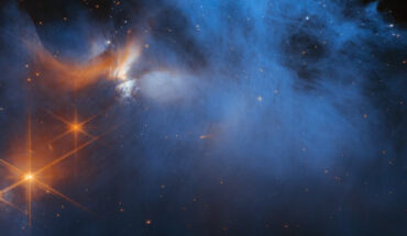This Webb image features the central region of the Chameleon I dark molecular cloud, which resides 630 light-years away in the constellation of Chamaeleon; the cold, wispy cloud material (blue, center) is illuminated in the infrared by the glow of the young, outflowing protostar Ced 110 IRS 4 (orange, upper left); the light from numerous background stars, seen as orange dots behind the cloud, can be used to detect ices in the cloud, which absorb the starlight passing through them. Image credit: NASA / ESA / CSA / M. Zamani, ESA & Webb / F. Sun, Steward Observatory / Z. Smith, Open University / Ice Age ERS Team.