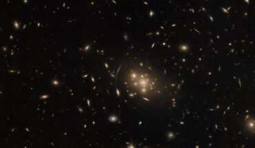 This Hubble image shows a large cluster of galaxies, SPT-CL J0019-2026, surrounded by various stars and smaller galaxies on a dark background. The color image was made from separate exposures taken in the visible and near-infrared regions of the spectrum with Hubble’s Advanced Camera for Surveys (ACS). It is based on data obtained through two filters. The color results from assigning different hues to each monochromatic image associated with an individual filter. Image credit: NASA / ESA / Hubble / H. Ebeling.