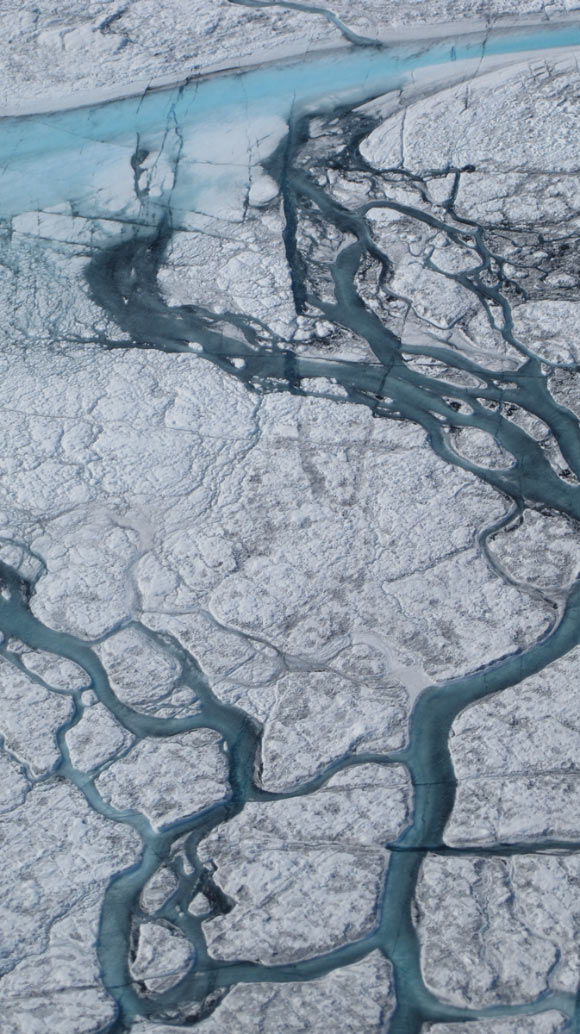 A large stream of meltwater (about 5 to 10 m in width) emerges from an upstream supraglacial lake in the Greenlandic ice on July, 21 2012. The darker shapes are minor streams covered by cryoconite (a mix of dust particles, soot, meteorite dust and organic material) which covers the ice sheet. Cracks cutting through the streams are visible. Along these cracks water can flow through, enlarging them and, eventually, generating moulins that can deliver meltwater through the ice. Image credit: M. Tedesco / CCNY / NASA ICE.