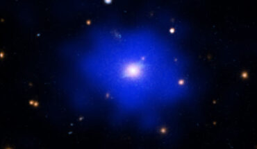 SPT-CL J2215-3537 resembles a blue cloud with a hazy golden light at its core. The cluster is set against the blackness of space, which is dotted with white, orange and blue specks of light. A combination of X-ray, ultraviolet, optical, and infrared light, was used to create the image. The blue cloud represents X-ray gas observed by NASA’s Chandra X-ray Observatory. The cloud is soft, and uniform in texture. Over time, galaxy clusters often merge or collide. These collisions cause disturbances and sharp features in a cluster’s gas. When a cluster takes on a smooth, calm appearance, as found here in SPT-CL J2215-3537, it indicates that no such collisions have occurred for a very long time. Astronomers call these galaxy clusters ‘relaxed.’ The hazy golden light at the heart of the cloud represents a massive central galaxy with a large amount of star formation. This central galaxy is quite isolated within the blue gas cloud. There are relatively few nearby specks of light near the central galaxy, and none are as bright or as large. No other large galaxies have been found within 600,000 light-years of the central galaxy. This implies that SPT-CL J2215-3537 has not experienced a galaxy merger in the last billion years. Image credit: NASA / CXC / MIT / M. Calzadilla / STScI / HST / N. Wolk.