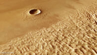 This oblique perspective view of Lycus Sulci and Yelwa Crater on Mars was generated from the digital terrain model and the nadir and color channels of the High Resolution Stereo Camera on ESA’s Mars Express. It shows the large 8-km-wide Yelwa Crater in the background, while the wrinkled terrain of Lycus Sulci dominates the foreground. These features lie on the edge of the ‘aureole’ of Olympus Mons. Image credit: ESA / DLR / FU Berlin / CC BY-SA 3.0 IGO.