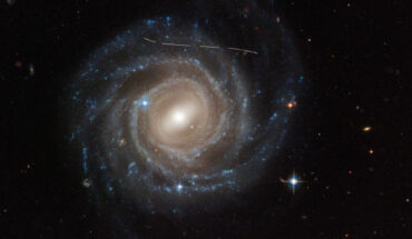 This Hubble image of the barred spiral galaxy UGC 12158 looks like someone took a white marking pen to it. In reality it is a combination of time exposures of a foreground asteroid moving through Hubble’s field-of-view, photobombing the observation of the galaxy. Several exposures of the galaxy were taken, what is evidence in the dashed pattern. The asteroid appears as a curved trail due to parallax: because Hubble is not stationary, but orbiting Earth, and this gives the illusion that the faint asteroid is swimming along a curved trajectory. The uncharted asteroid is in inside the asteroid belt in our Solar System, and hence is 10 trillion times closer to Hubble than the background galaxy. Image credit: NASA / ESA / Hubble / Pablo García Martín, UAM / Joseph DePasquale, STScI / Alex Filippenko, UC Berkeley.