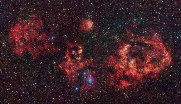 This VST image shows a cloud of gas and dust, shaped like a cosmic bat. The intense red glow comes from hydrogen atoms ionized by the intense radiation of young stars within the cloud. The most prominent objects here are RCW 94, which represents the right wing of the bat, and RCW 95, which forms the body, while the other parts of the bat have no official designation. Image credit: ESO / VPHAS+ Team / VVV Team.