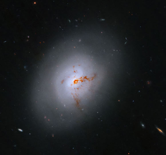 This Hubble image reveals the lenticular galaxy NGC 1266; reddish-brown clumps and filaments of dust partially obscure the galaxy’s full face, while red, blue, and orange light from distant galaxies shines through its diffuse outer regions and dots the inky-black background. Image credit: NASA / ESA / K. Alatalo, STScI / G. Kober, NASA & Catholic University of America.